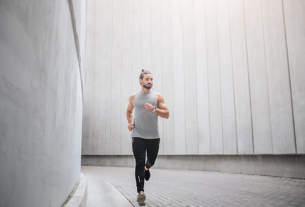 Zdjęcie joggera biegnie przez betonowy korytarz. Macha rękami i patrzy w prawo. Poważny młody człowiek jest skoncentrowany.