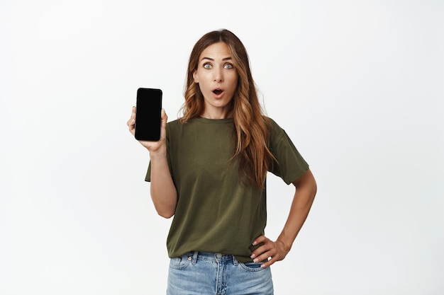 Zdjęcie kobiety pokazującej ekran telefonu komórkowego ze zdziwioną, zachwyconą twarzą, sprawdzającą nową aplikację, pokazującą interfejs aplikacji ze zdumionym wyrazem twarzy, białe tło.