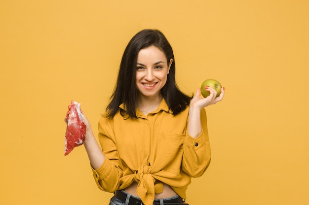 Zdjęcie koncepcyjne słodkiej samicy wybiera między mięsem a jabłkiem. Nosi żółtą koszulę, na białym tle żółty kolor tła.