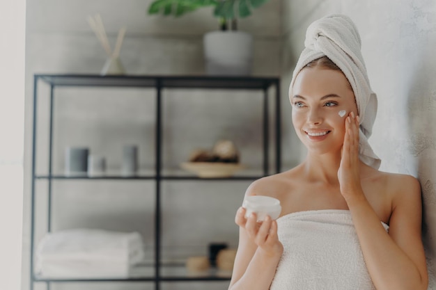 Zdjęcie Ładna kobieta nakłada krem do twarzy zawinięty w stojaki na ręczniki w łazience Koncepcja pielęgnacji skóry
