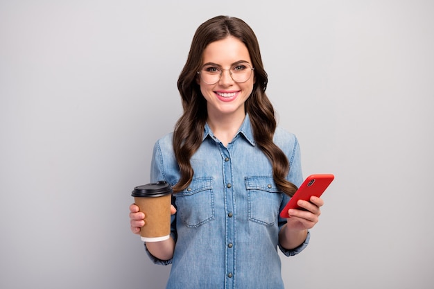 Zdjęcie ładnej freelancerki pani trzymaj kubek papierowy gorąca kawa napój przerwa w pracy przeglądanie telefonu urządzenie nosić specyfikacje na co dzień dżinsy dżinsowa koszula na białym tle szary kolor