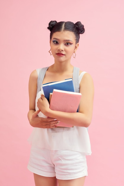 Zdjęcie ładnej studentki w wieku 20 lat z fryzurą z podwójnymi bułkami, noszącej plecak, trzymającej wiele książek do nauki izolowanych na różowym tle