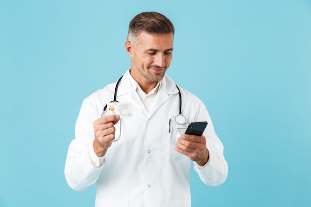 Zdjęcie lekarza w średnim wieku na sobie biały fartuch i stetoskop trzymając smartfon, stojąc na białym tle nad niebieską ścianą