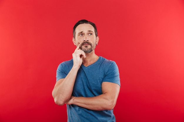 Zdjęcie mężczyzny w wieku 30 lat rozmyślające spojrzenie w swobodnej koszulce, patrząc w górę i myśląc lub pamiętając, na białym tle na czerwonym tle