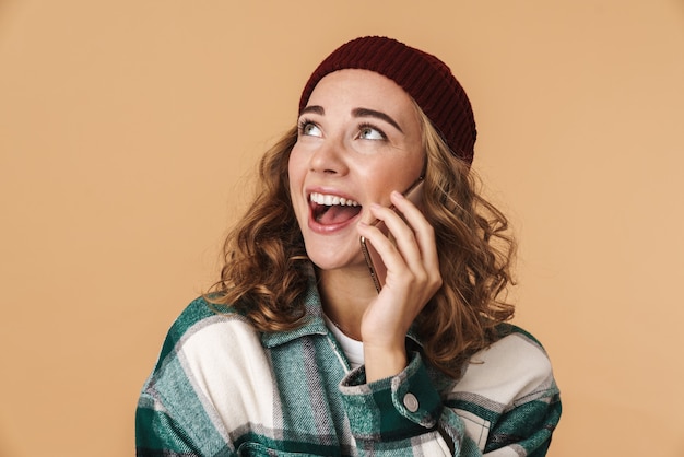 Zdjęcie miłej, wesołej kobiety w czapce z dzianiny, uśmiechającej się i rozmawiającej przez telefon komórkowy na beżowym tle