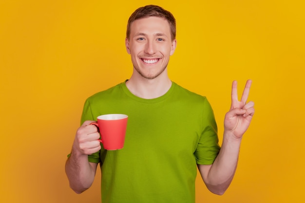 Zdjęcie zdjęcie młodego człowieka szczęśliwy pozytywny uśmiech napój filiżanka kawy pokaż pokój fajny znak v na białym tle nad żółtym kolorem tła