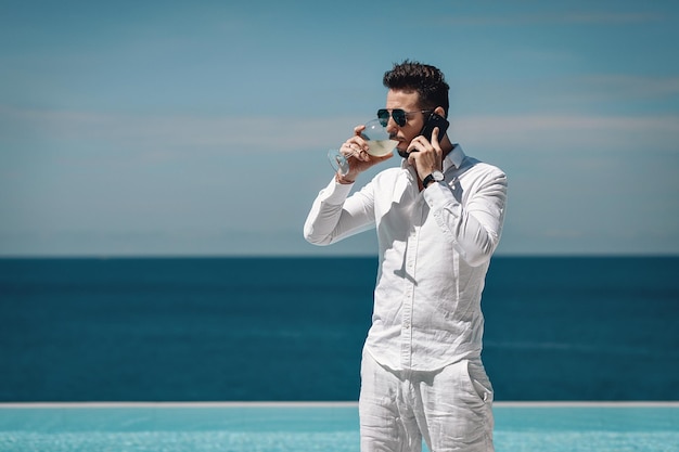 Zdjęcie młodego człowieka w białej koszuli stoi przy basenie i rozmawia przez telefon komórkowy, pijąc mojito i ciesząc się pięknym widokiem na błękitne morze. Koncepcja podróży i biznesu. Phuket. Tajlandia.