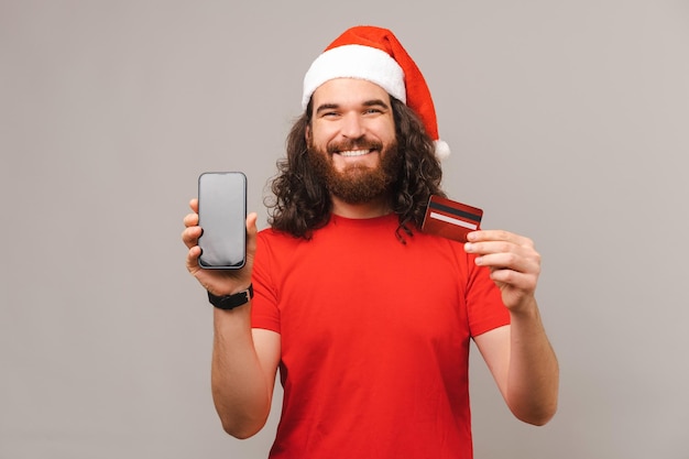 Zdjęcie młodego szczęśliwego brodatego mężczyzny w kapeluszu Świętego Mikołaja i pokazującym ekran karty kredytowej i smartfona