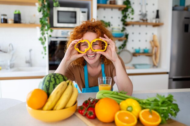 Zdjęcie młodej kobiety uśmiecha się i trzyma kółka pieprzu na oczach podczas gotowania sałatki ze świeżymi warzywami w kuchni w domu