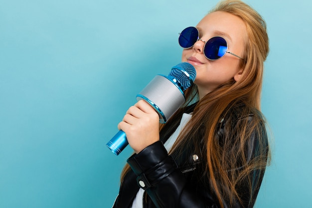 Zdjęcie nastolatki w niebieskich okularach przeciwsłonecznych na niebieskiej ścianie, dziewczyny z mikrofonem w bujanej czarnej kurtce