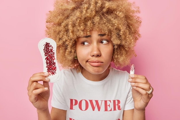 Zdjęcie niezadowolonej kobiety z kręconymi włosami opowiada o krwawieniu podczas menstruacji trzyma brudną podpaskę higieniczną i tampon pokazuje różne rodzaje kobiecych produktów higienicznych na różowej ścianie