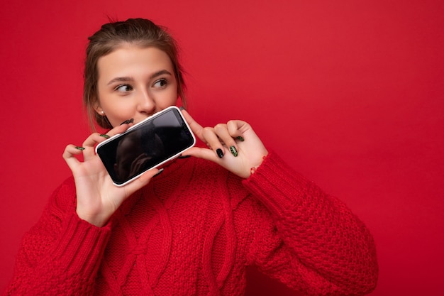 Zdjęcie piękne słodkie młoda kobieta sobie ciepły czerwony sweter na białym tle nad czerwonym tle ściany trzymając smartfon i pokazując telefon z pustym wyświetlaczem na makieta patrząc z boku.