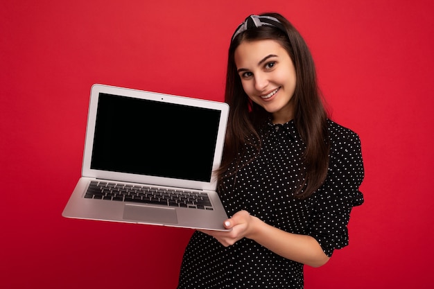 Zdjęcie Piękne Uśmiechnięte Ciemne Włosy Dziewczyny Na Sobie Czarne Ubrania Trzymając Netbook Patrząc Na Kamery Na Białym Tle Na Tle Kolorowe ściany. Pusty Ekran Monitora, Makieta