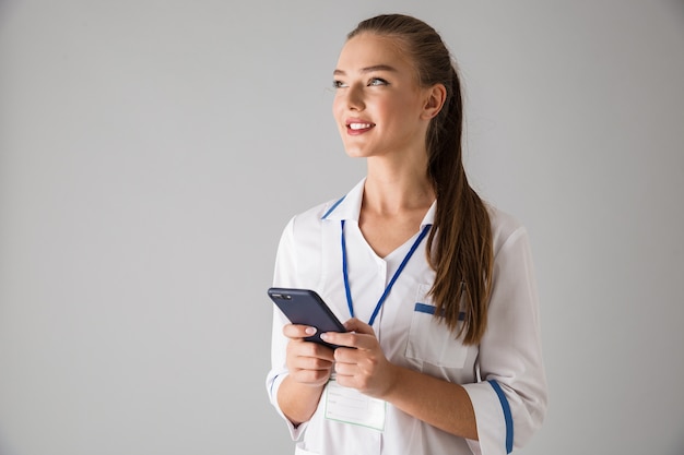 Zdjęcie pięknej szczęśliwej młodej kobiety lekarz kosmetolog na białym tle na szarej ścianie trzymając telefon komórkowy za pomocą.