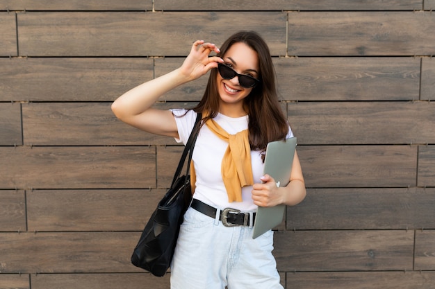 Zdjęcie pięknej uśmiechniętej młodej damy ubrana w białą koszulkę jasnoniebieskie dżinsy i żółty sweter trzymający komputer przenośny i czarne okulary przeciwsłoneczne, pozostając na ulicy i patrząc na kamery