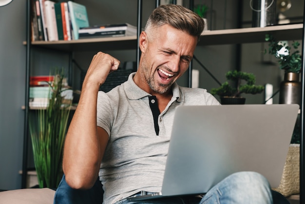 Zdjęcie podekscytowanego dorosłego mężczyzny w swobodnym ubraniu, uśmiechającego się i świętującego podczas korzystania z laptopa w mieszkaniu