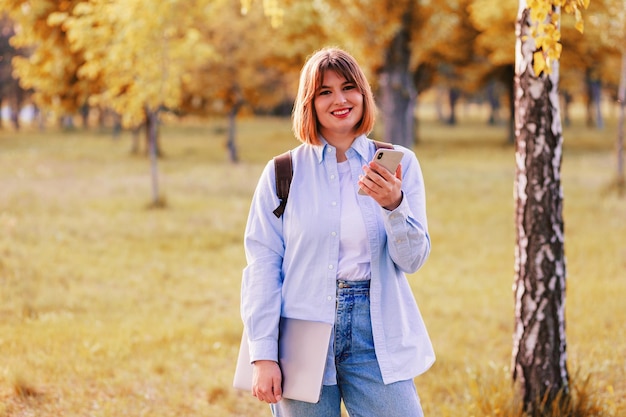 Zdjęcie portret studentki trzymającej telefon i laptopa i uśmiechającej się na zewnątrz w parku
