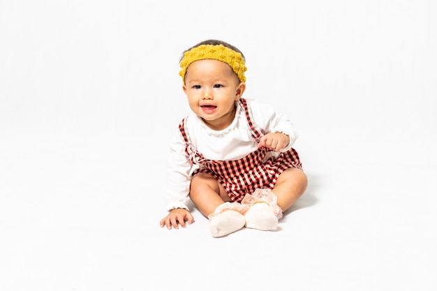 Zdjęcie Portretowe Małego Dziecka, W Wieku 9-10 Miesięcy, Azjatycka Dziewczyna Ubrana W Białą Koszulę Z Kolorowym Kombinezonem I żółtym Paskiem Do Włosów, Siedząca Na Białym Tle.