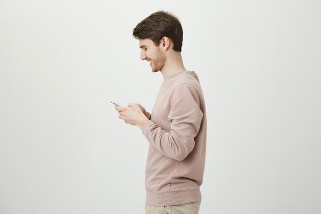 Zdjęcie profilowe atrakcyjnego mężczyzny z modną fryzurą i włosiem, trzymającego smartfona i śmiejącego się z czegoś, co widzi na ekranie