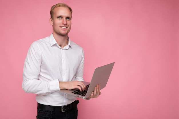Zdjęcie profilowe po stronie strzał przystojny uśmiechający się pewnie blond dorosły mężczyzna osoba posiadająca komputer laptop pisania na klawiaturze na sobie białą koszulę patrząc na kamery na białym tle na różowym tle.