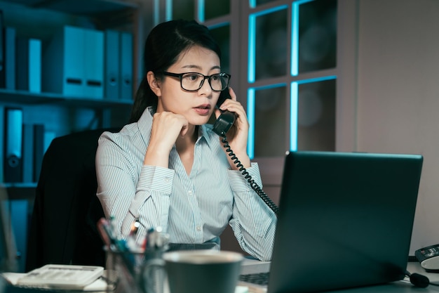 Zdjęcie przekonana młoda businesswoman pracy na komputerze przenośnym podczas rozmowy przez telefon. właściwa pracownica patrząca na monitor komputera przenośnego i dyskutująca z partnerem korporacyjnym na telefon w nocy