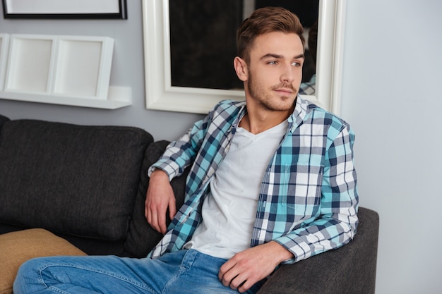 Zdjęcie przystojny młody mężczyzna ubrany w koszulę w klatce wydruku i dżinsy, siedząc na kanapie w domu i patrząc na bok.