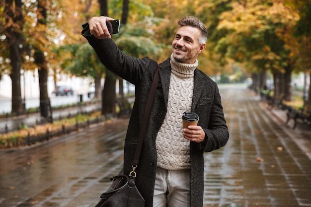 Zdjęcie radosnego mężczyzny w wieku 30 lat w ciepłym ubraniu spacerującego po jesiennym parku na świeżym powietrzu i robiącego selfie na telefonie komórkowym