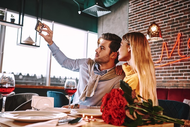 Zdjęcie romantycznej kolacji w drogiej restauracji. Młoda para uśmiecha się i robi zdjęcie selfie na telefonie podczas kolacji