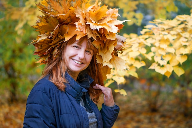Zdjęcie ślicznej dojrzałej kobiety uśmiechającej się do aparatu, wieniec z żółtych liści klonu, pożółkłe opadające liście, spacer i piękny niezapomniany zapach jesieni, miejskiego parku, na ulicy