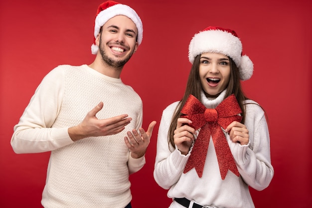 Zdjęcie zdjęcie słodkiej pary w świątecznych kapeluszach, zadowolony z wakacji. mężczyzna i kobieta są zakochani, wyglądają błogosławieni i uśmiechają się, odizolowane na czerwonym tle.