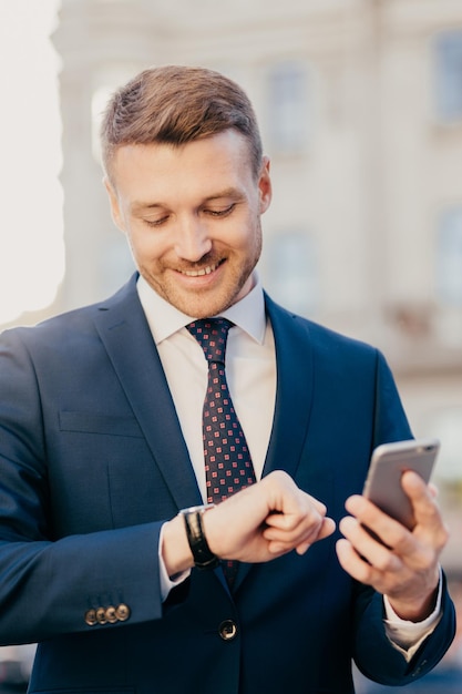 Zdjęcie sprytnego męskiego prawnika czeka na klienta na zewnątrz patrzy na zegarek trzyma inteligentny telefon, czekając na ważne połączenie, nosi formalny garnitur, ma przyjemny uśmiech na twarzy Koncepcja spotkania ludzi w czasie