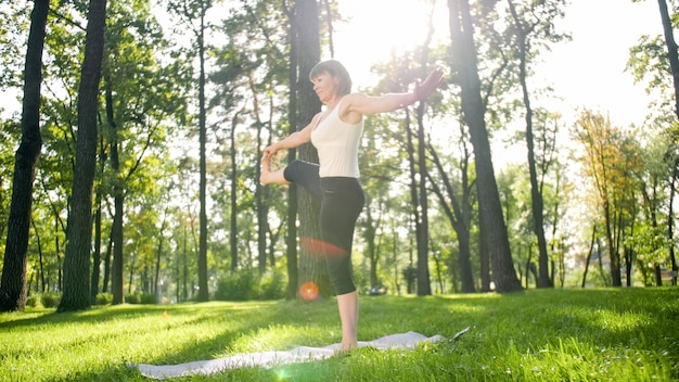 Zdjęcie średniej wieku uśmiechnięta kobieta praktykuje jogę i medytuje w parku. Kobieta rozciągająca się i ćwicząca fitness na macie w lesie