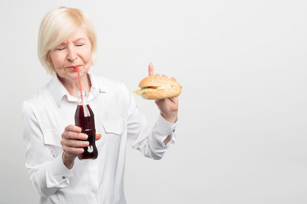 Zdjęcie starej kobiety degustującej coca-colę i jedzącej burgera. Nie lubi zdrowego stylu życia. Woli jeść smaczne, ale tuczące i złe jedzenie.