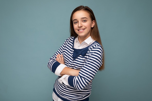 Zdjęcie strzał pięknej szczęśliwej uśmiechniętej brunetki małej dziewczynki noszącej pasiasty longsleeve stojącej na białym tle nad niebieskim tle ściany patrząc na kamerę