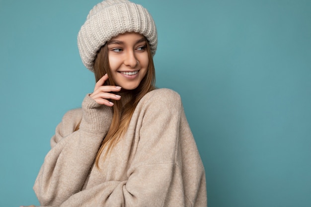 Zdjęcie Strzał Piękny Szczęśliwy Uśmiechający Się Seksowny Młody Ciemny Blond Kobieta Na Białym Tle Nad Niebieskim Tle ściany Sobie Beżowy Ciepły Sweter I Beżowy Kapelusz Z Dzianiny, Patrząc Z Boku