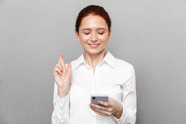 Zdjęcie szczęśliwa młoda ładna ruda biznesowa kobieta pozowanie na białym tle nad szarą ścianą za pomocą telefonu komórkowego zrobić nadzieję, że palce skrzyżowane gest.