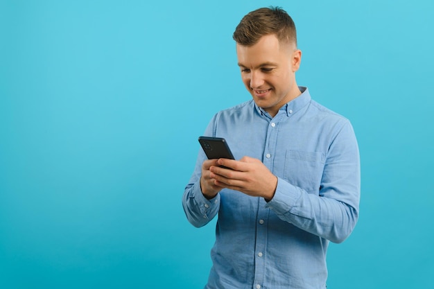 Zdjęcie szczęśliwego przystojnego mężczyzny trzymającego telefon komórkowy w ręku wysyłającego SMS-y Młody facet z dużym uśmiechem toothy pozuje na niebieskim tle