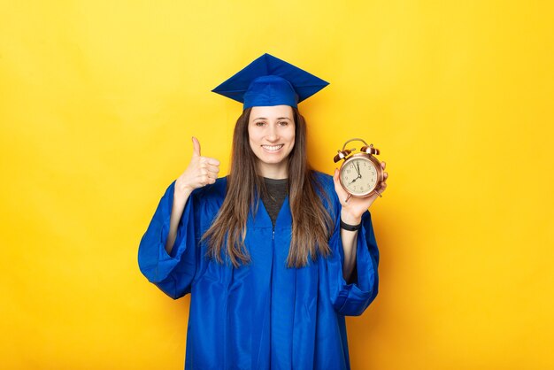 Zdjęcie Szczęśliwej Kobiety Kończącej Studia Pokazując Kciuk Do Góry I Budzik Na żółtym Tle