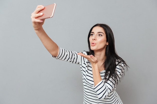 Zdjęcie szczęśliwej młodej kobiety wyślij pocałunek podczas robienia selfie na szarej powierzchni. Spójrz na telefon.