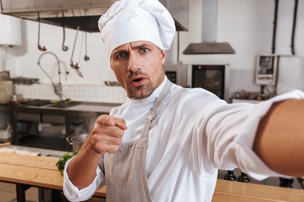 Zdjęcie szczęśliwy wódz mężczyzna w fartuchu, biorąc selfie, stojąc w kuchni w restauracji