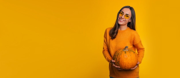 Zdjęcie transparentu ślicznej uśmiechniętej młodej dziewczyny w okularach z dojrzałą dynią w rękach, podczas gdy ona pozuje na białym tle na żółtym tle w sezonie jesiennym