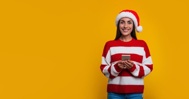Zdjęcie transparentu szczęśliwej pięknej kobiety w kapeluszu Świętego Mikołaja z inteligentnym telefonem w rękach, używającego go i patrzącego z uśmiechem na aparat