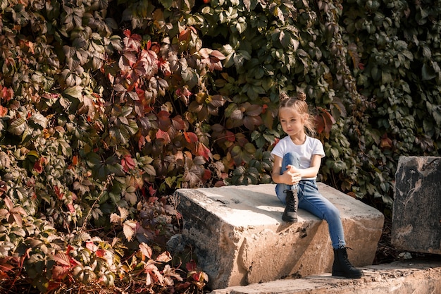 Zdjęcie Uliczne Małej Pięknej Dziewczyny W Dżinsach I Białej Koszulce Na Tle Betonowych Płyt I Jesiennych Liści