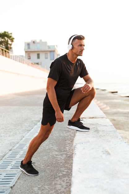 Zdjęcie umięśnionego mężczyzny w wieku 30 lat w krótkich spodenkach i koszulce podczas treningu nad morzem i słuchania muzyki przez słuchawki bezprzewodowe podczas wschodu słońca