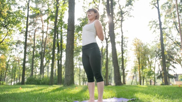 Zdjęcie Uśmiechnięta Szczęśliwa Kobieta 40 Lat Robi ćwiczenia Jogi Na Macie Fitness W Lesie. Harmonia Człowieka W Naturze. Osoby W średnim Wieku Dbające O Zdrowie Psychiczne I Fizyczne