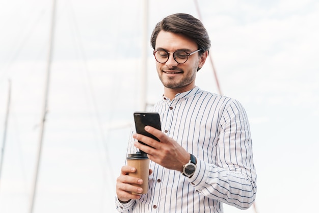 Zdjęcie uśmiechniętego młodego mężczyzny w okularach, piszącego na telefonie komórkowym i pijącego kawę z papierowego kubka podczas spaceru po molo