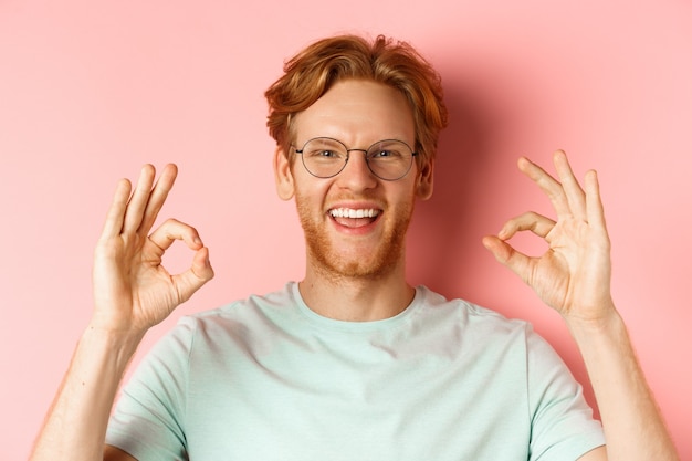 Zdjęcie zdjęcie w głowę portret atrakcyjnego młodzieńca z rozczochranymi rudymi włosami i brodą, w okularach, uśmiechającego się z białymi zębami i pokazującego aprobatę znaków ok, chwalcie coś dobrego.