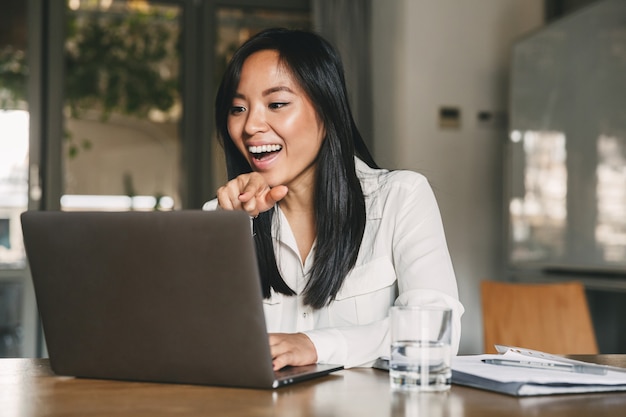 Zdjęcie wesołej Azjatki lat 20. w białej koszuli, śmiejącej się i wskazującej palcem na ekranie laptopa, podczas rozmowy lub rozmowy wideo w biurze