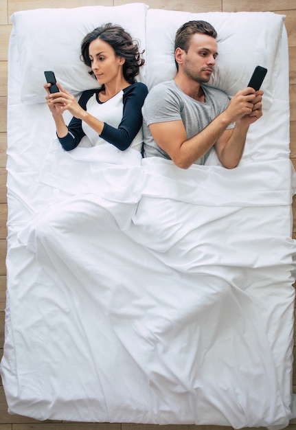Zdjęcie z góry przedstawiające piękną kobietę i przystojnego młodzieńca korzystających ze smartfonów, leżąc w dużym białym łóżku. Gadać na czacie online, rozmawiać online. Powyższe zdjęcie pary.