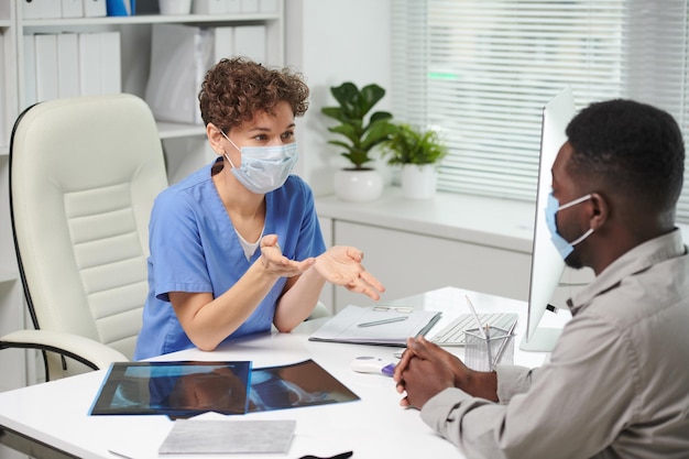 Zdjęcie z wysokim kątem widzenia profesjonalnego pracownika medycznego w niebieskim mundurze siedzącego przed pacjentem opowiadającego o problemach zdrowotnych